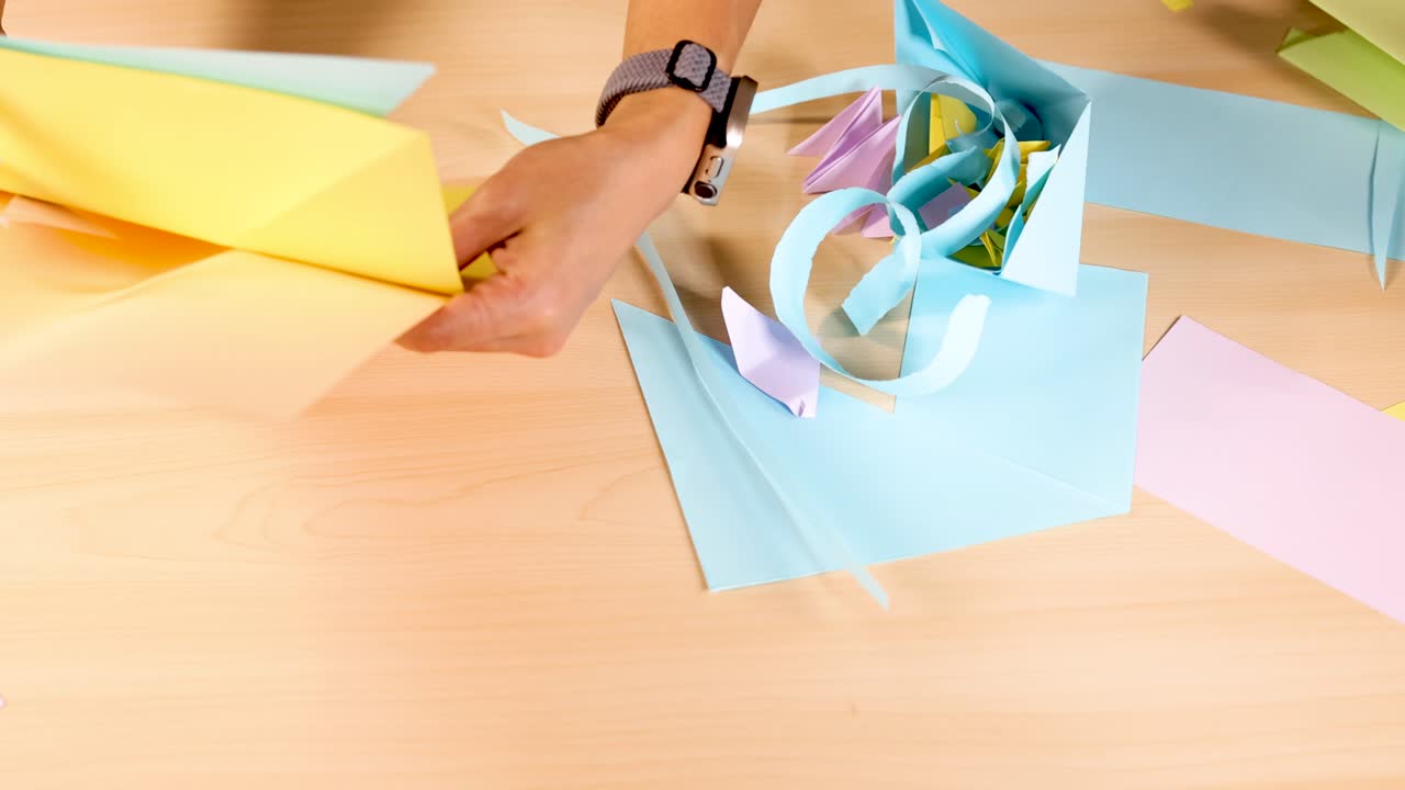 Hands fold pastel origami paper under bright lighting, overhead view, with paper crafts visible