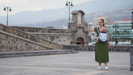 While-strolling-with-her-baby-in-a-kangaroo-backpack,-a-young-woman-documents-their-travels-using-a-mobile-phone.-She-captures-moments-and-looks-at-the-screen