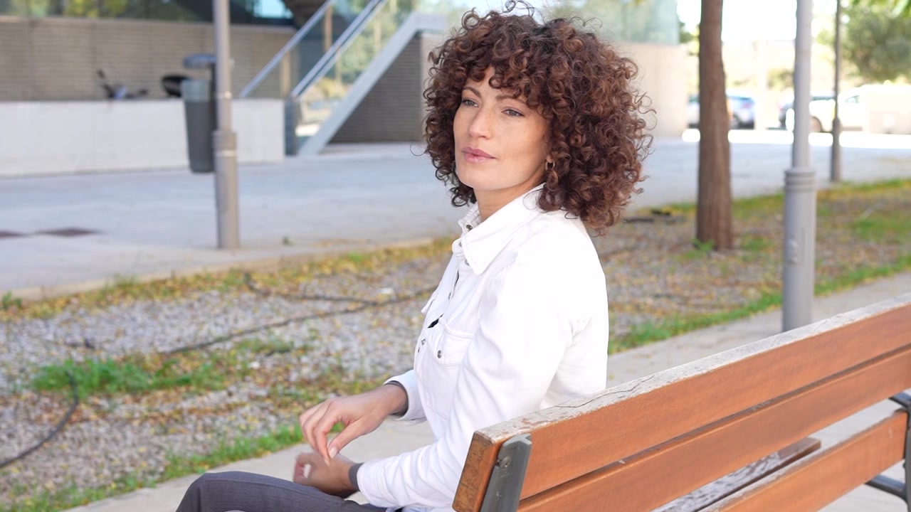 Una giovane donna pensierosa che indossa un abbigliamento elegante è seduta su una panchina del parco di giorno.
