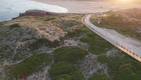 Aerial-flyover-view-of-motorhome-parked-on-Bordeira,-Portugal-coastal-hillside-overlooking-sunlit-beach-and-ocean-waves