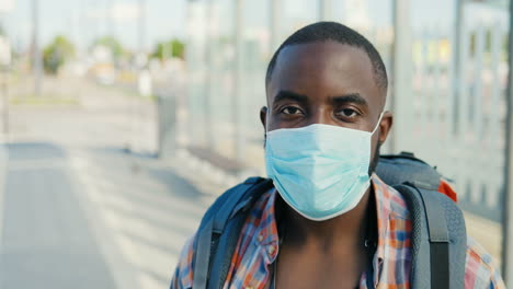 retrato de un joven viajero afroamericano con mochila y máscara médica mirando la cámara al aire libre
