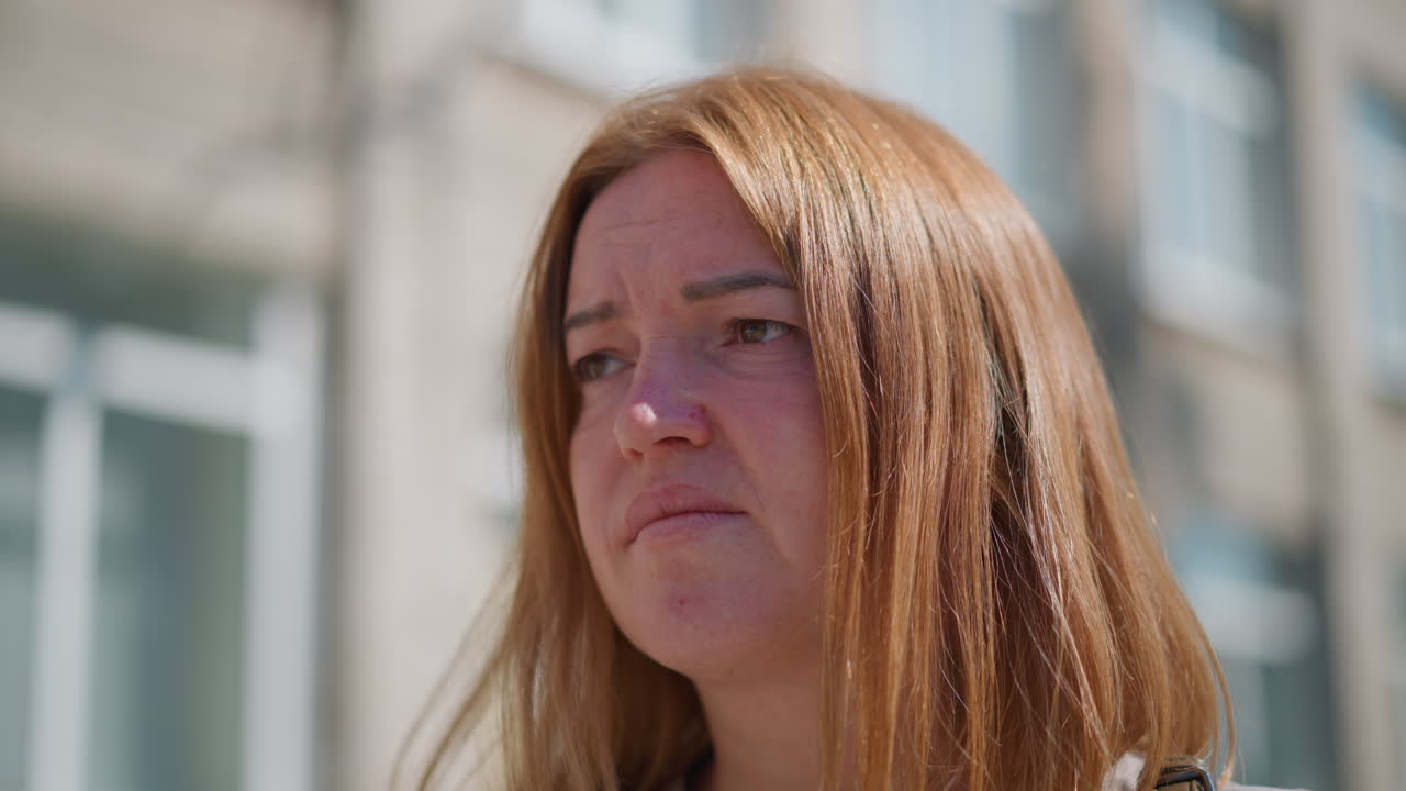 Primo piano di una donna preoccupata con i capelli lunghi che guarda pensierosa verso il basso sotto la luce del sole, con una canottiera, sfondo sfocato di un edificio residenziale