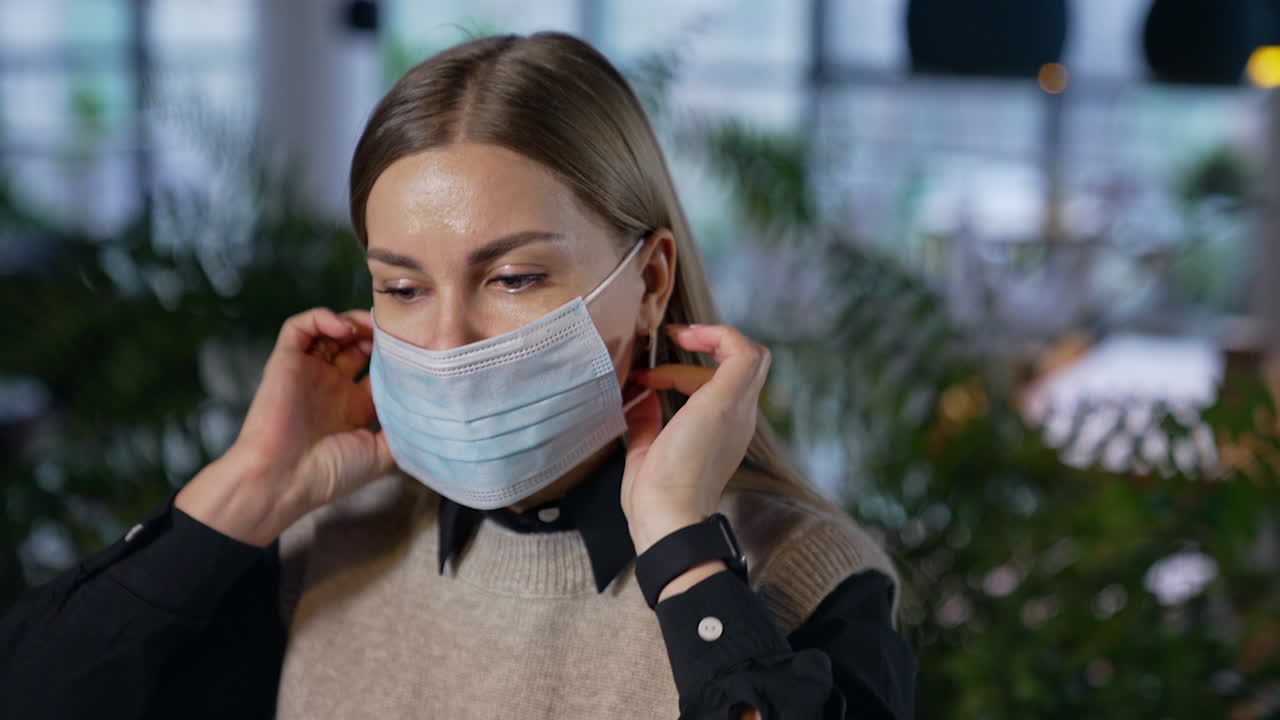 Attractive female office worker puts on mask. Lady in mask looks straight into the camera. Woman in mask against the blurred backdrop.
