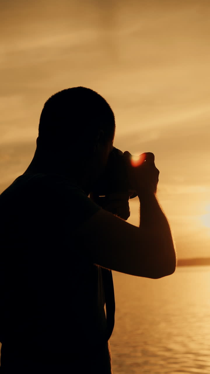 A man with a camera in his hands is photographing the sunset by the river on the background of people bathing. Close-up Vertical video