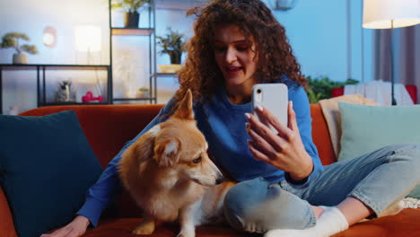 Woman-pet-lover-kissing-cute-corgi-dog-and-taking-selfie-via-smartphone-on-sofa-at-home-living-room