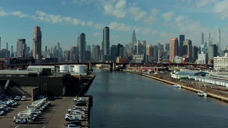 Landing-footage-of-Newtown-Creek-waterfront.-Cars-on-fenced-parking-lot-and-industrial-site-behind.-Skyline-with-skyscrapers.-New-York-City,-USA