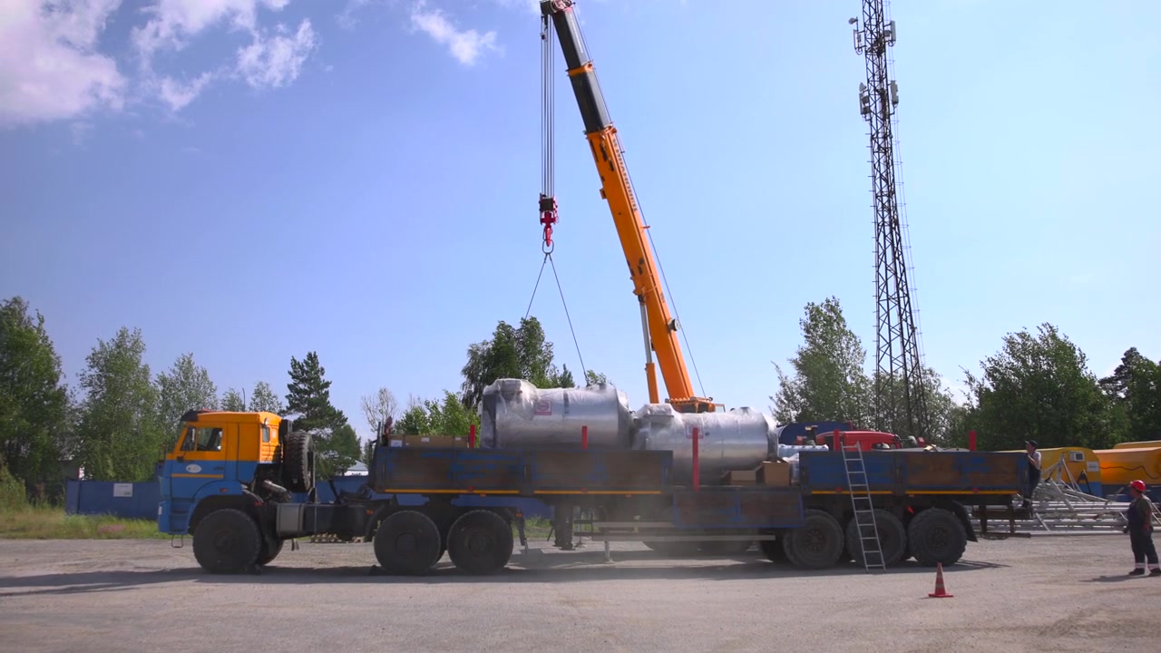 Orange mobilkran med udskudt bom løfter rød krog og stropper over en ladvogn, der transporterer store indpakkede cylindriske tanke, mens to arbejdere i sikkerhedsudstyr koordinerer i nærheden på en udendørs læsseplads.