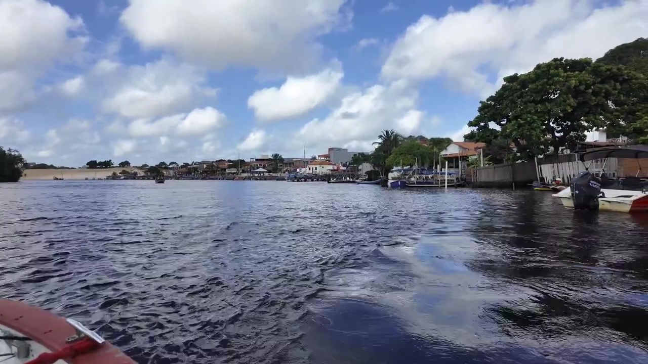 Rekaman POV dari haluan perahu yang bergerak melintasi Sungai Preguiça yang lebar, menuju tepi sungai kota Barreirinhas di bawah langit biru cerah penuh awan di Maranhão, Brasil