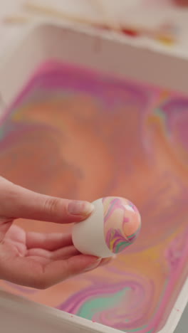 lady shows easter egg with abstract pattern of ebru art over tray with color mix at table closeup slow motion. spring holiday traditions. creative lifestyle