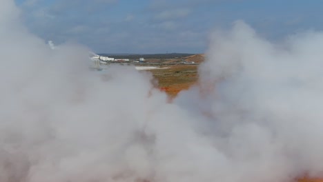 aerial past steaming hot fumeroles in a geothermal aera reveals a geothermal power plant in the distance