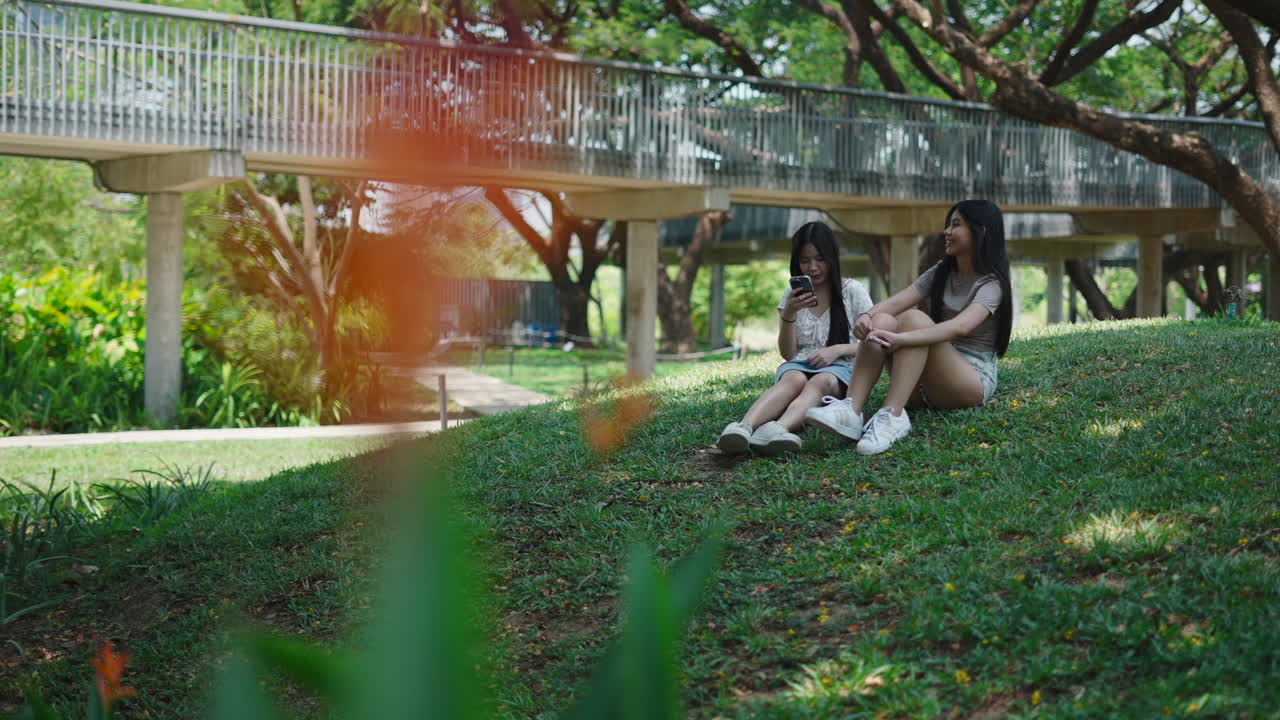 Dos mujeres asiáticas jóvenes sentadas charlando en una colina cubierta de hierba en un parque