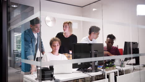 creative business team gathered around a laptop computer in a small office cubicle, seen through glass