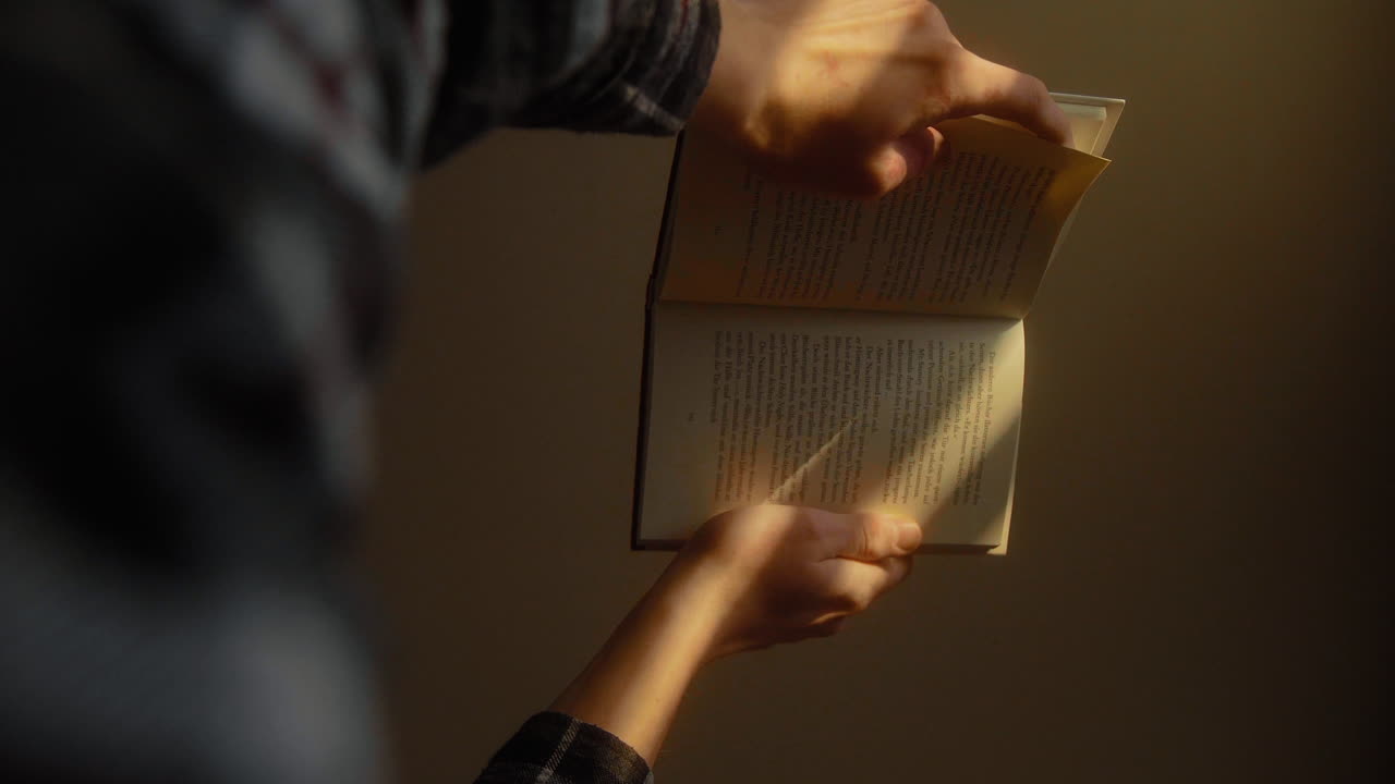 Young male holding a book in his hands reading in sunlit living room at home relaxing. Close Up 4K Lookin Up