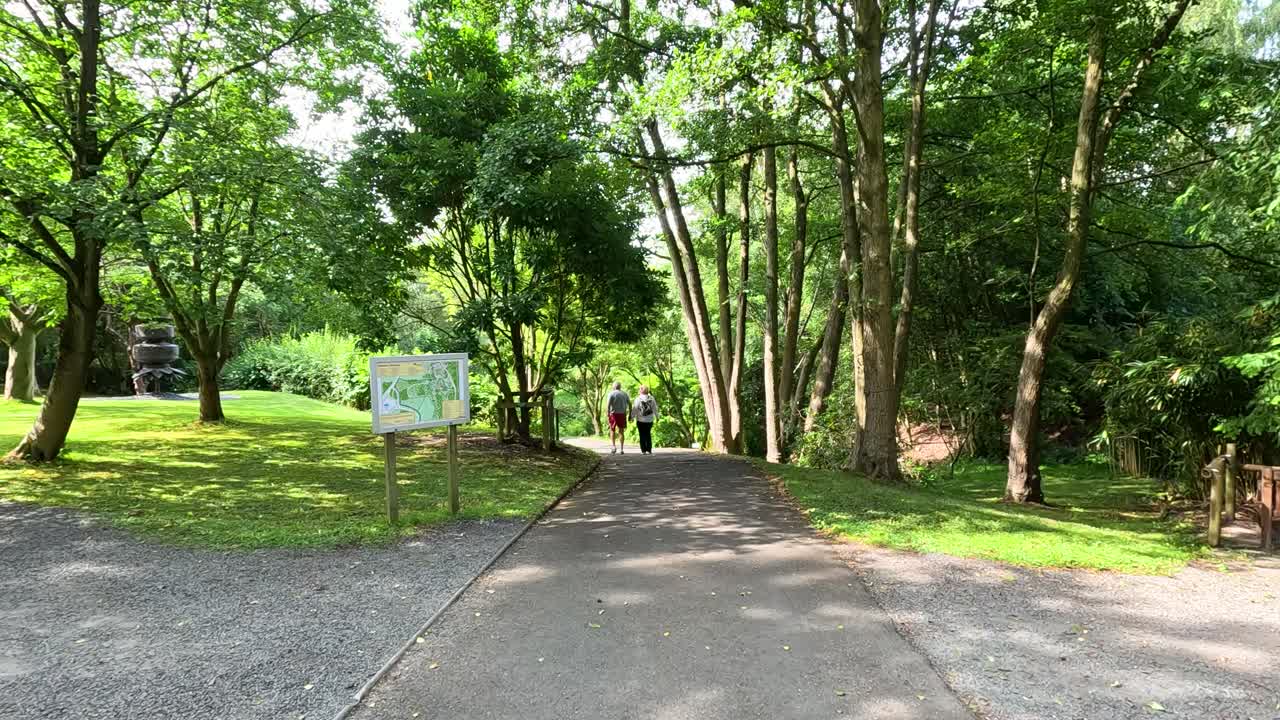 Two people stroll down a paved path through lush, sunlit botanical gardens, surrounded by green trees and informational signs, with steady forward camera movement