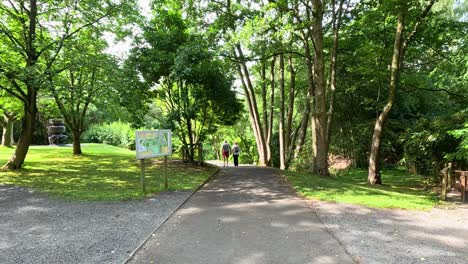 two people stroll down a paved path through lush, sunlit botanical gardens, surrounded by green trees and informational signs, with steady forward camera movement