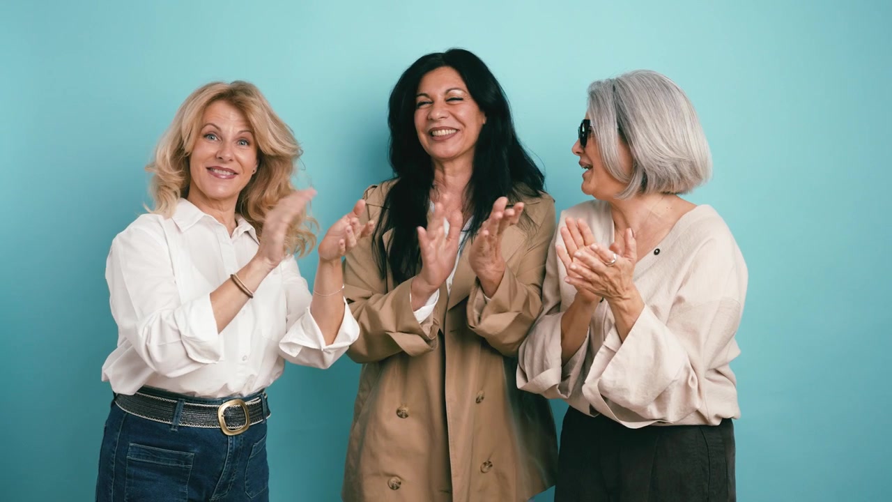 Three Mature Women Smiling and Clapping Together Against a Blue Background