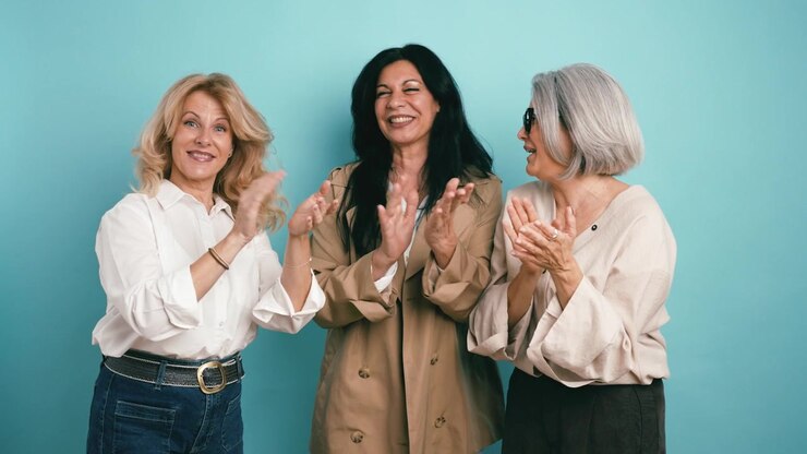 Tre donne mature che sorridono e applaudono insieme su sfondo blu