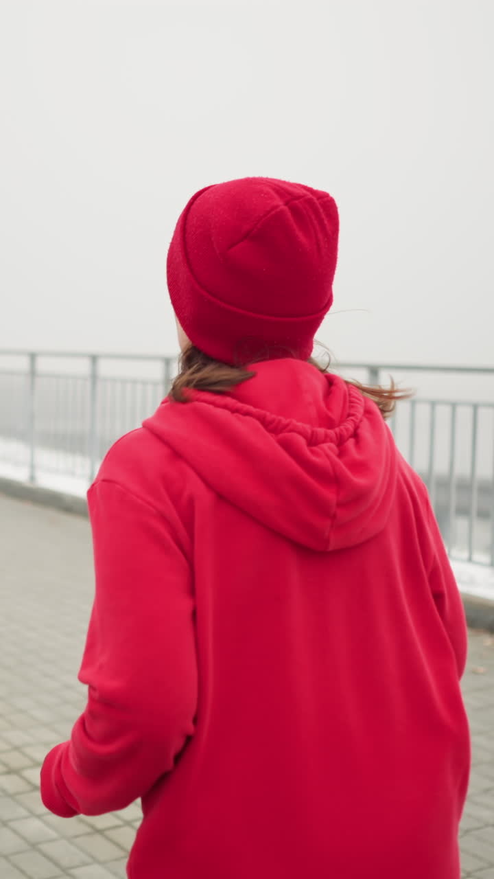 woman in red beanie and hoodie jogging along foggy pathway near iron railing with distant bridge and moving cars visible breath rising amid frosty trees capturing active fitness