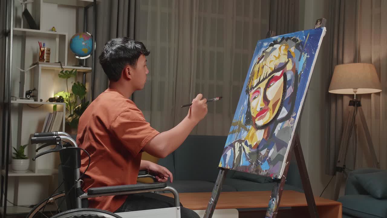 Asian Artist Boy In Wheelchair Holding Paintbrush And Mixing It With Colour Before Painting A Girl'S Face On The Canvas