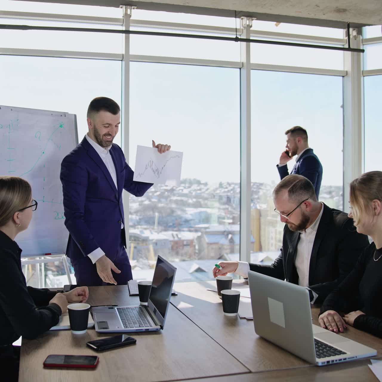 Businessman holding a paper with graph on it and showing it to his colleagues. Coworkers sitting at the table and looking at the paper. Man in a suit standing at the window and talking on the phone at the backdrop