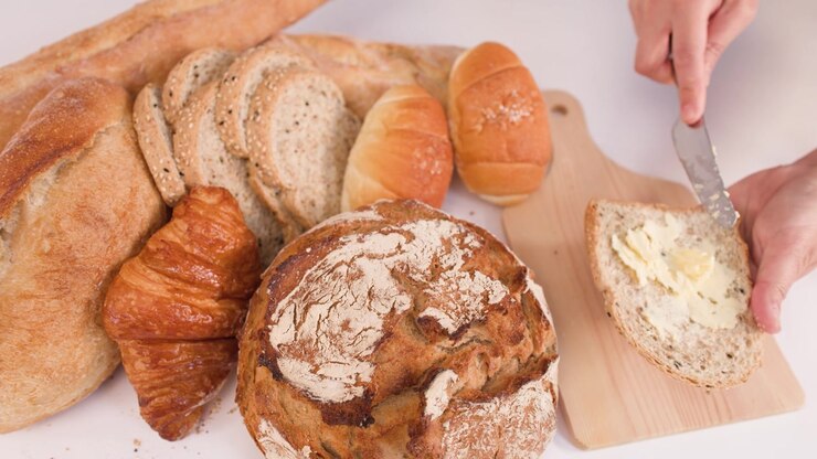 Hands use a knife to spread creamy butter on multigrain bread beside various fresh loaves.