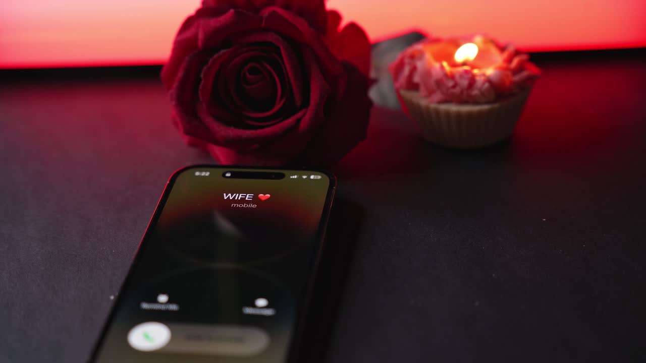 Incoming call from “Wife” on smartphone lying on dark table with red rose and lit candle, glowing red ambient light and soft bokeh, romantic evening communication concept