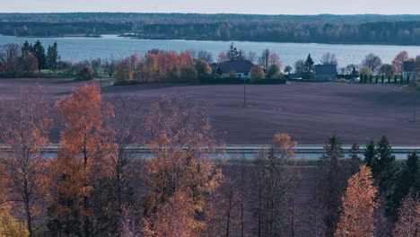 Bird's-eye-view-of-a-countryside-road-with-beautiful-autumn-colored-trees,-houses,-and-a-river-in-the-background