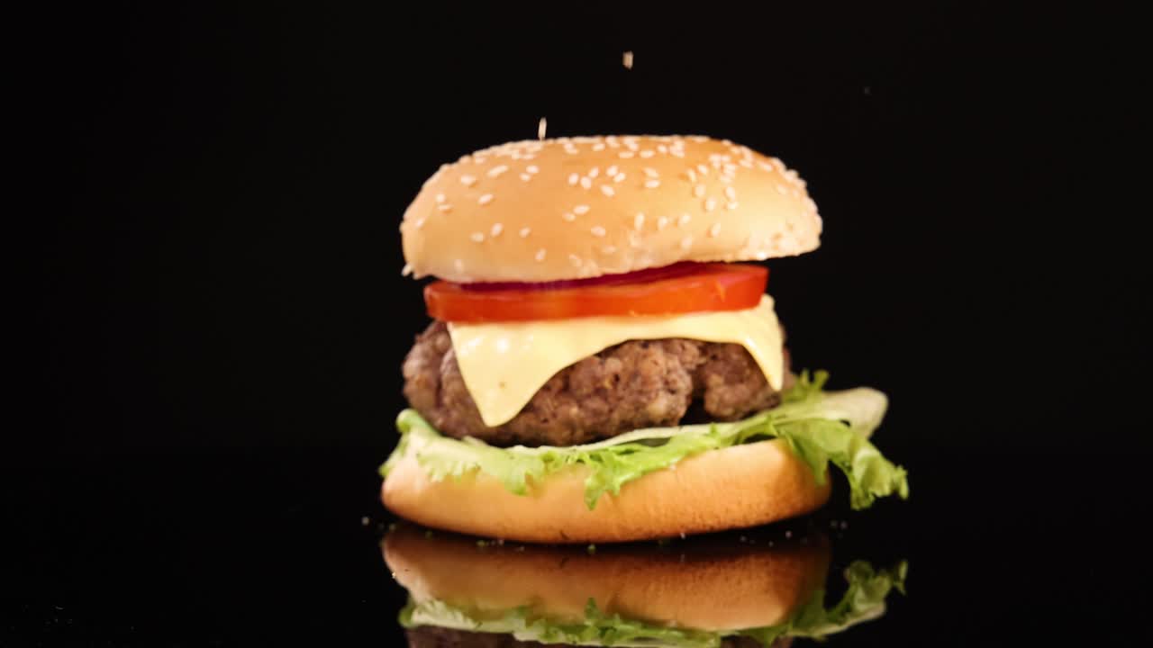 A golden onion ring falls in slow motion onto a cheeseburger with lettuce and tomato, surrounded by onion rings, against a black background with dramatic lighting