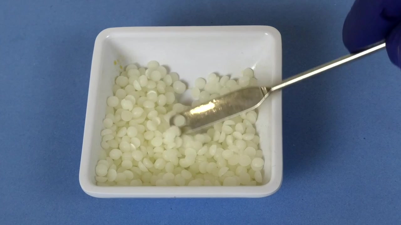 Pharmacien(ne) avec des gants en nitrile pesant des granulés de cire blanche à l'aide d'une spatule en laboratoire.