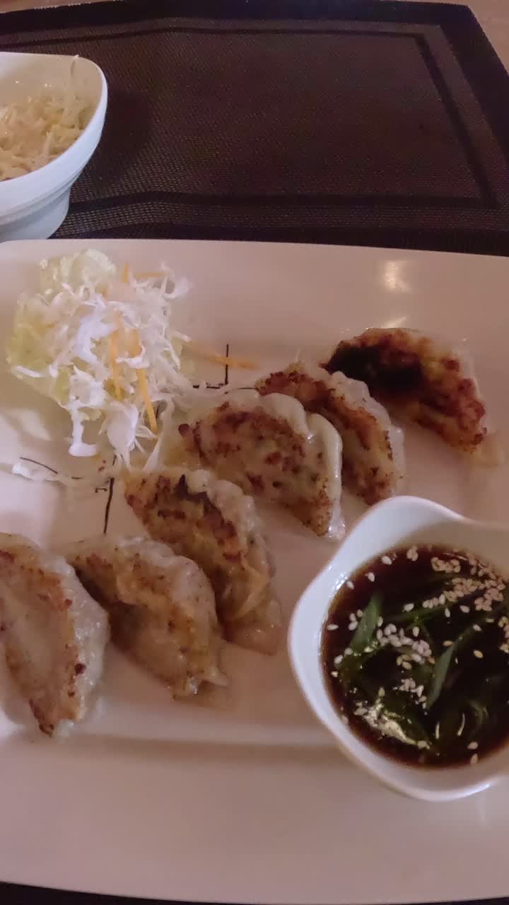 Overhead view, hand using chopsticks to pick and dip a piece of fried gyoza in soy sauce with sesame seeds