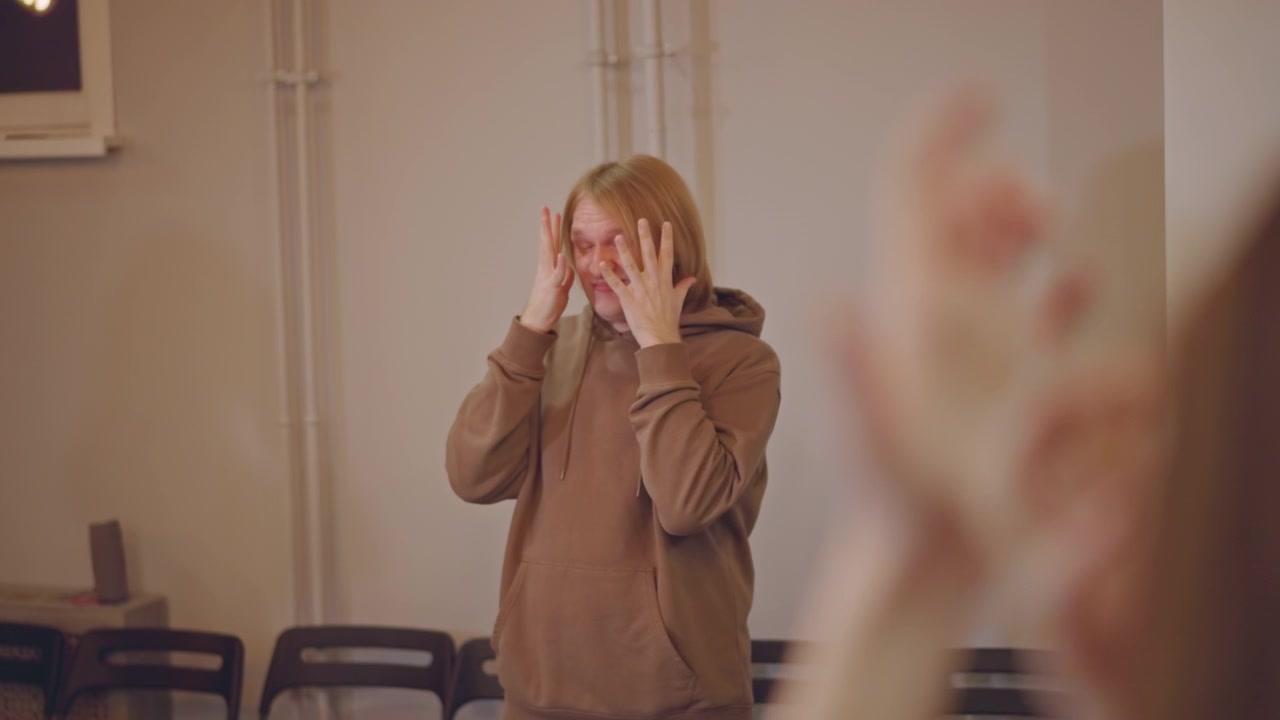 Caucasian teen singer warming up breath in rehearsal room, hoodie and stacked chairs visible, hands near head showing nervous concentration, soft warm lighting, coach figure in foreground