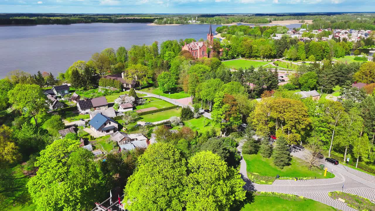 A red brick church rises from a dense green canopy in a riverside village, surrounded by scattered cottages and fields under a vivid spring sky in rural Europe.