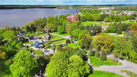a red brick church rises from a dense green canopy in a riverside village, surrounded by scattered cottages and fields under a vivid spring sky in rural europe.