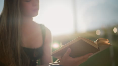 sun rays reflect off book held by woman seated outdoors, legs crossed, as wind gently blows through pages, background is softly blurred