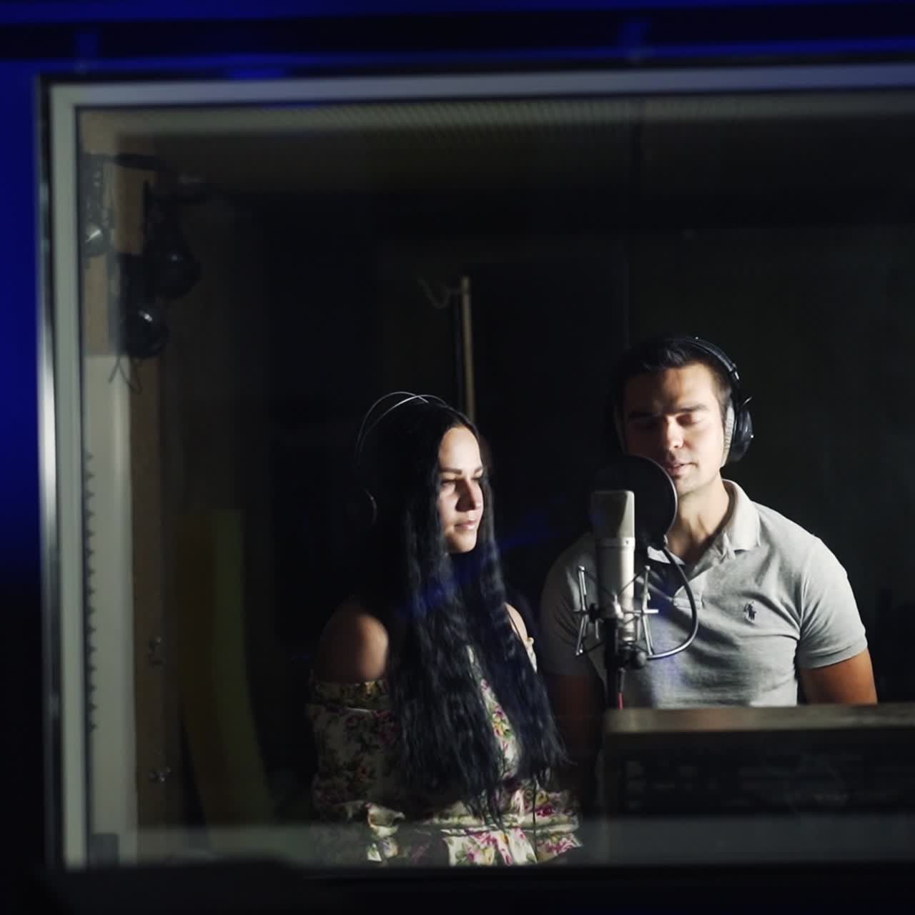 Equipment with a screen working in the music recording studio. A couple wearing headphones stand in front of microphone next room behind window