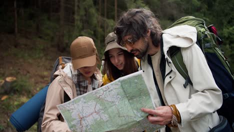 a trio of hikers in trekking attire with large backpacks survey their path on a large paper map they hold in their hands. orientation in the area during the travell