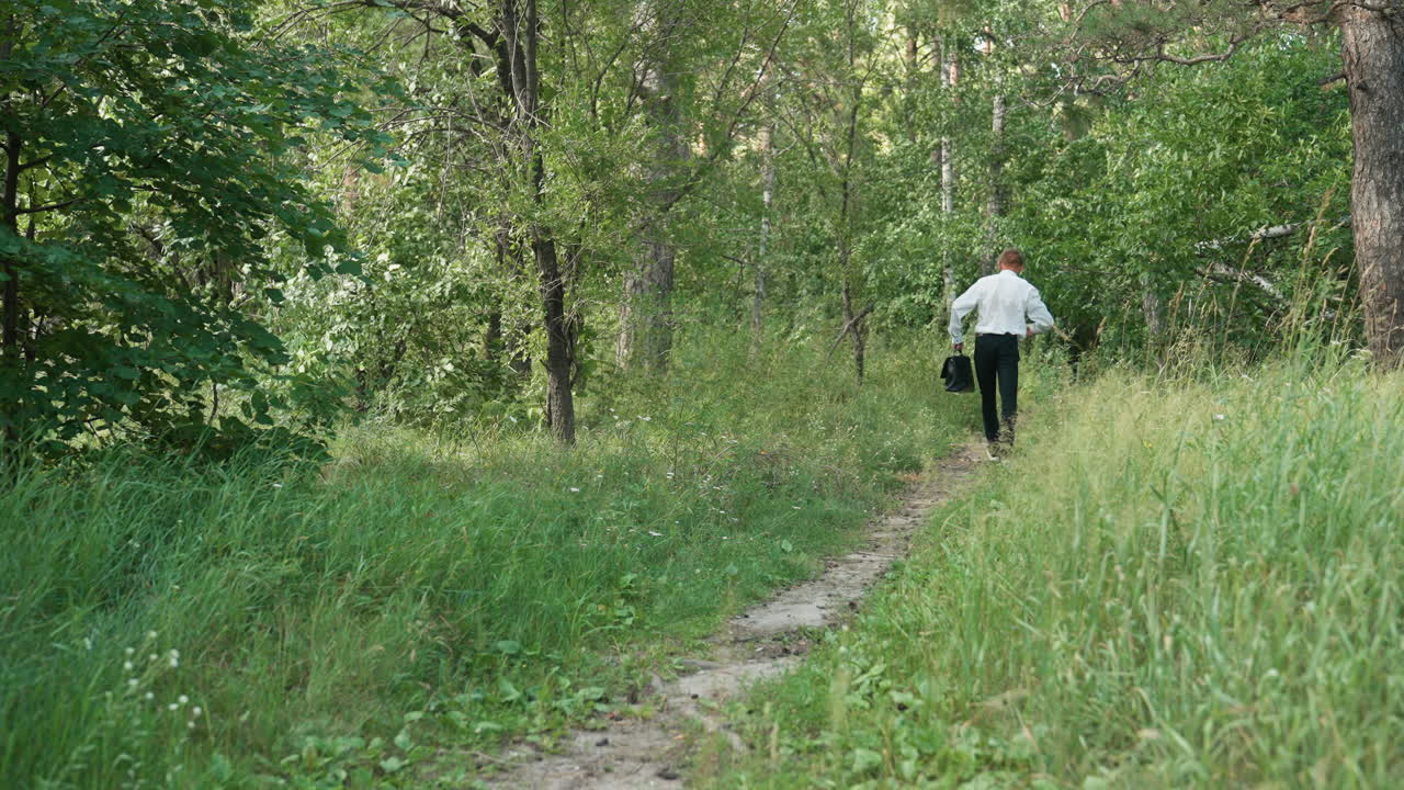 Back view of botany student in white shirt and black trousers carrying backpack walking slowly into forest path, pointing hands and observing plants along way surrounded by lush greenery