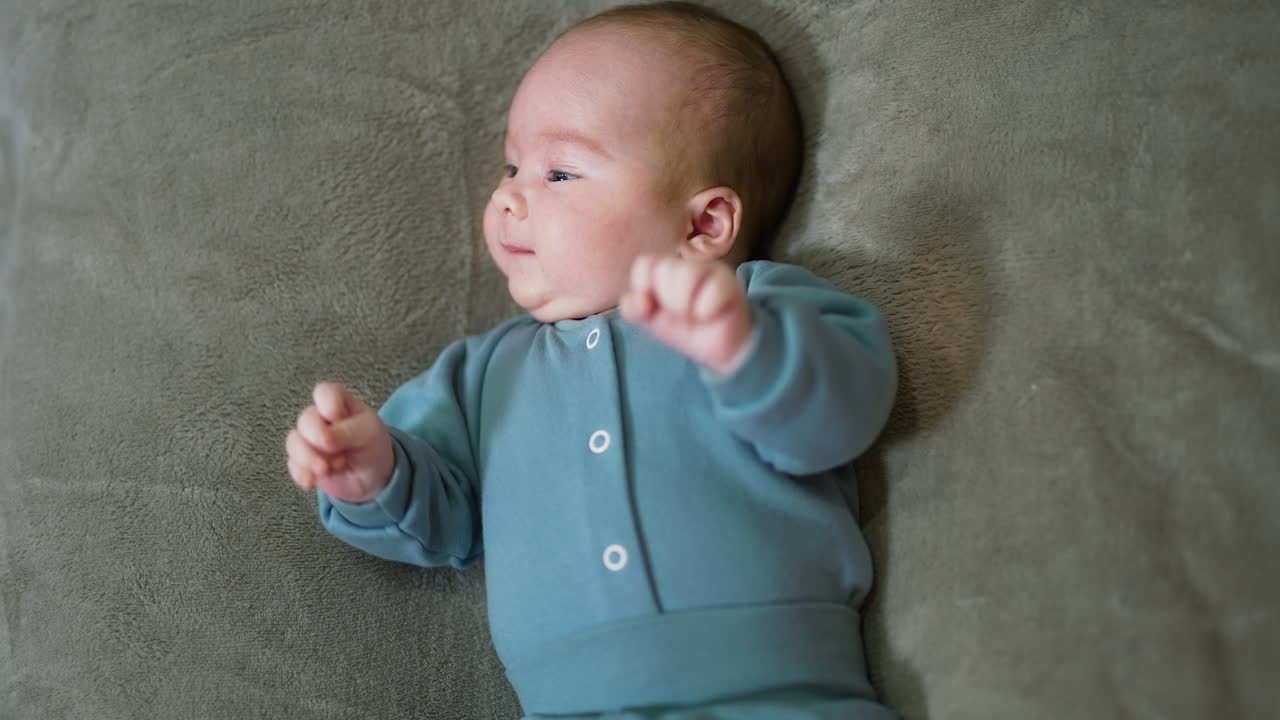 Cute newborn baby boy lies on the bed and looks aside. Adorable kid in blue clothes moves his arms actively. Top view.