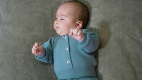 cute newborn baby boy lies on the bed and looks aside. adorable kid in blue clothes moves his arms actively. top view.