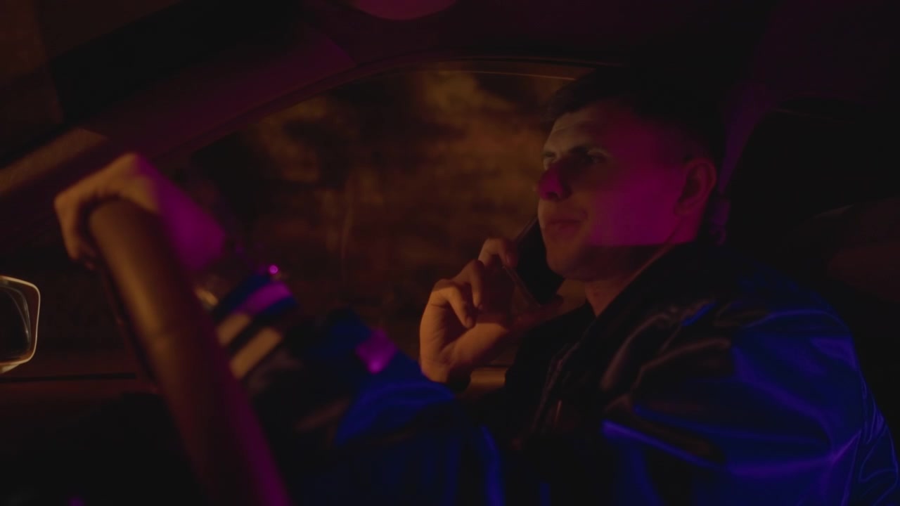 Guidatore bianco in auto di notte, telefono all'orecchio, sguardo contemplativo, giubbotto di pelle, bagliore del cruscotto, riflessi magenta e ambrati, riflessi dei lampioni urbani, atmosfera calma e cinematografica, turno di notte solitario