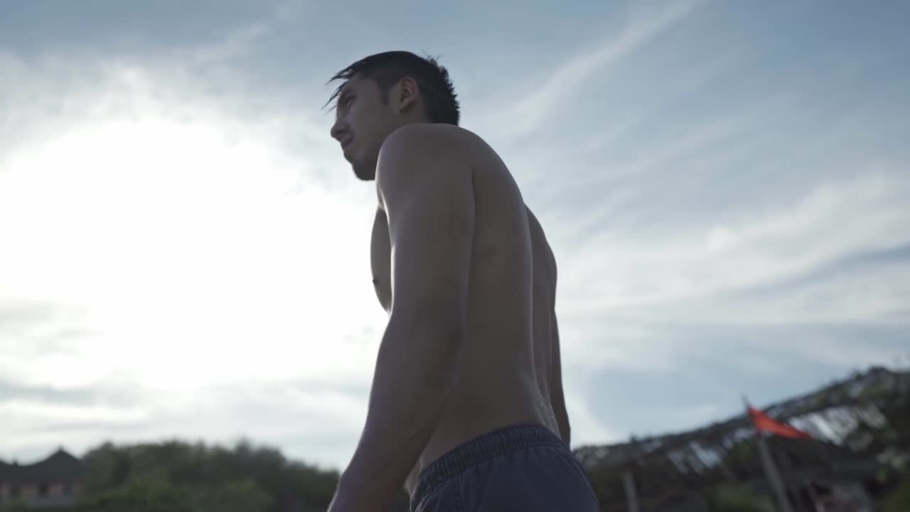 A low-angle shot captures a man in swim shorts walking away from the camera and along the water's edge on a sandy beach in Zipolite, México