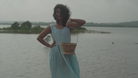 slow motion static shot of a charming young woman in light blue dress with black hair standing by the lake with a wood basket in her hand in windy weather