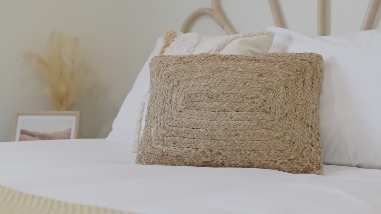 Movimento di camera circolare al rallentatore che mostra un cuscino in juta naturale con manici su biancheria da letto bianca e pulita in un ambiente luminoso e minimalista di una camera da letto