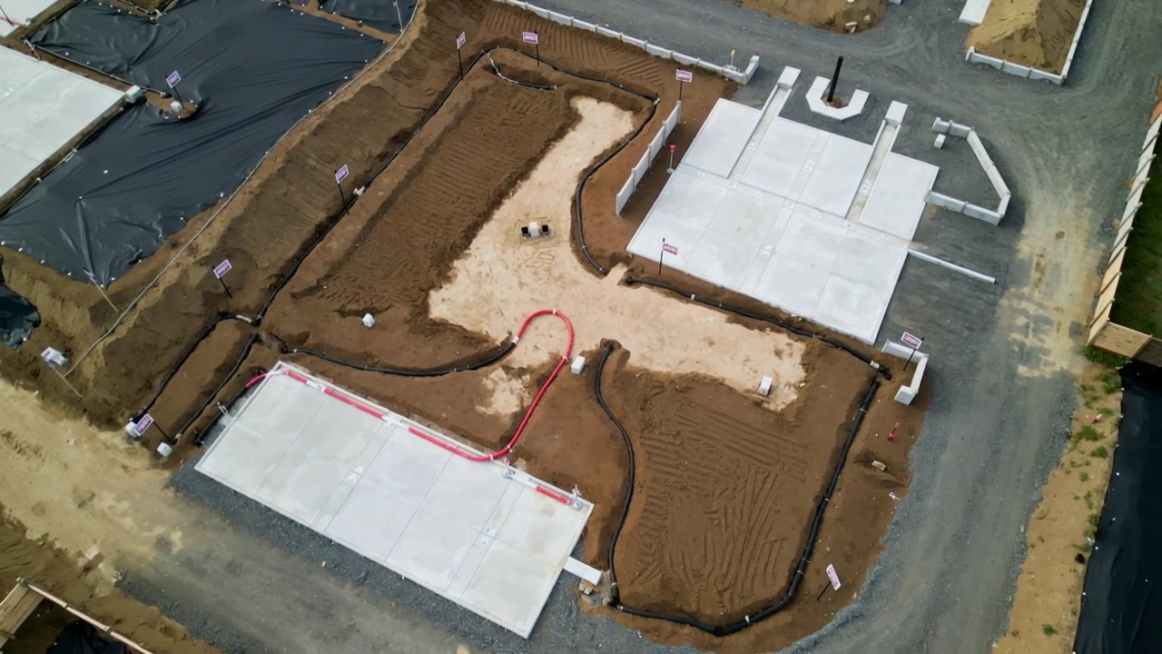 Drone che si avvicina e si inclina su un lotto residenziale, mostrando piazzole, una barriera antisabbia e un anello di condotto rosso.