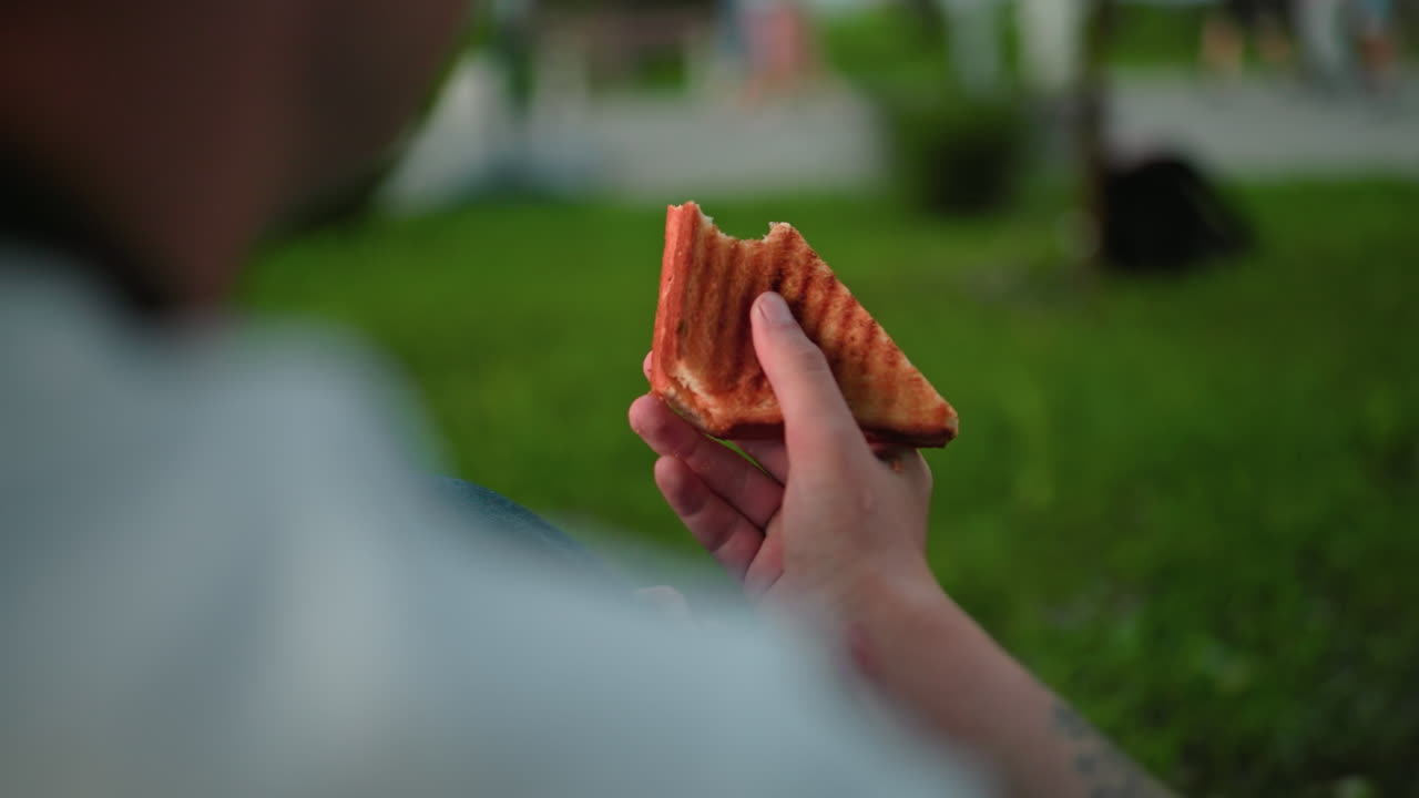 vista trasera de cerca de alguien con una camiseta blanca sosteniendo un sándwich medio comido observándolo con personas y vegetación borrosa en el fondo