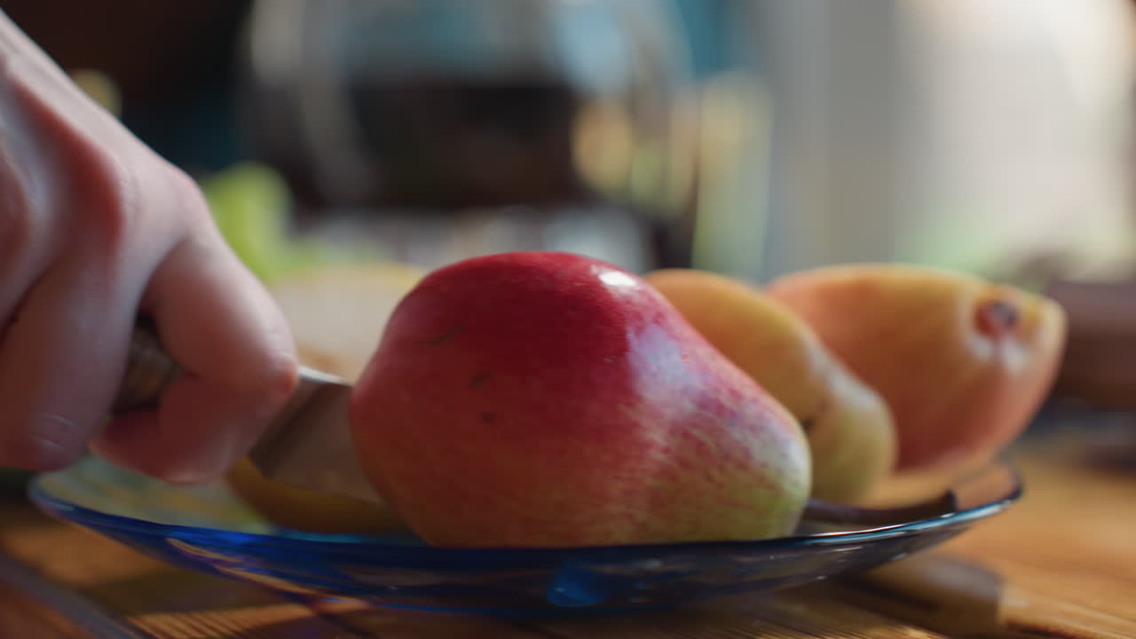 Close up of person slicing fresh apple with sharp knife on glass plate, pear and green grapes visible in background, scene set on wooden table under warm natural light in cozy indoor environment