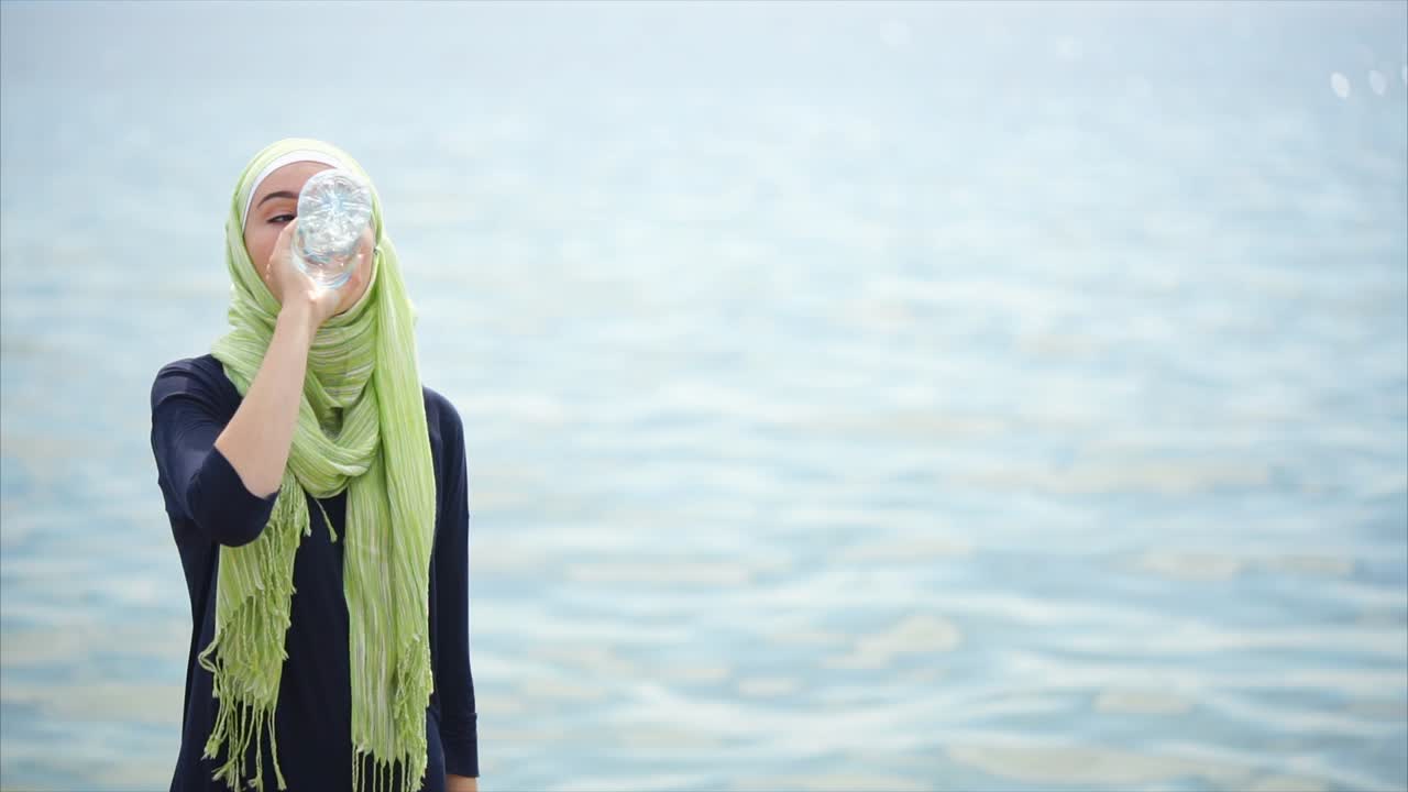 Woman in Hijab Drinking Water by the Ocean