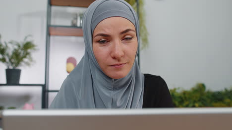 a muslim woman in hijab smiles at the camera while working on her laptop in a home office setting.