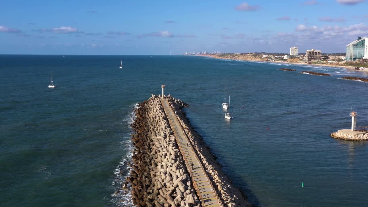 Multiple sailing yachts with folded sails en route to the marina of Herzeliya in Israel navigating between the breakwater and the red buoy on the calm Mediterranean. Wide drone dolly shot