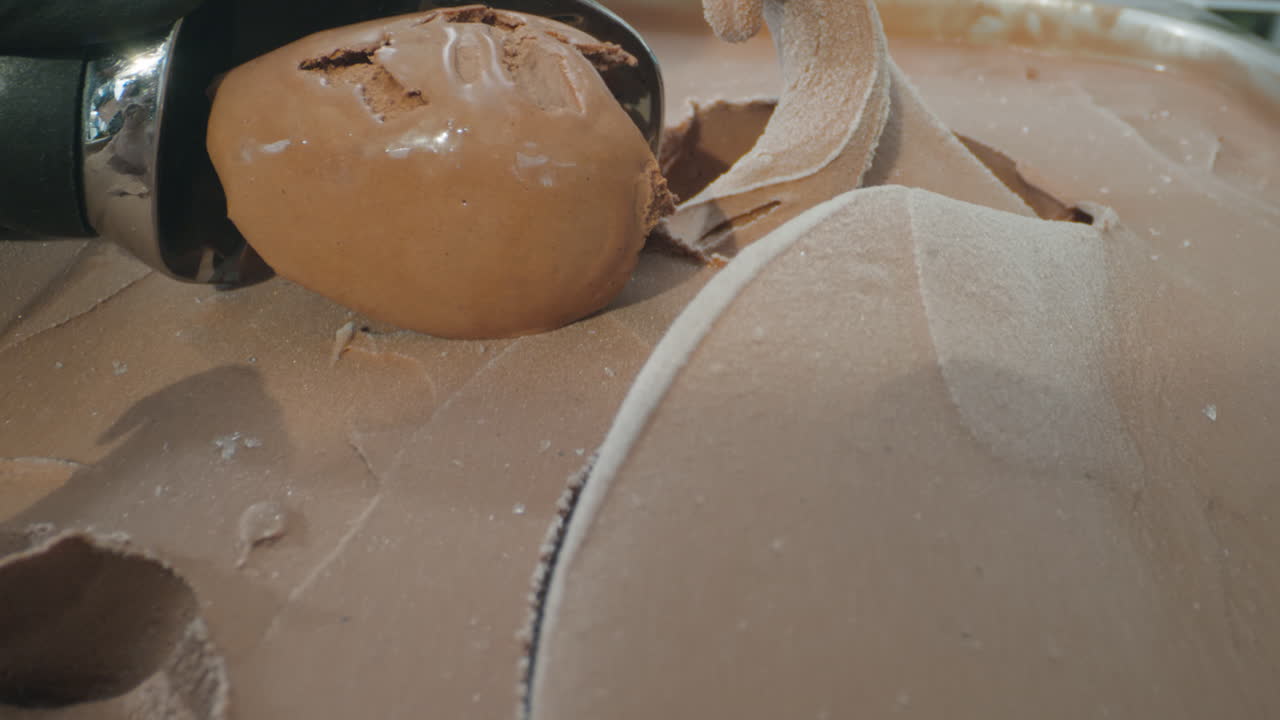 Close-up slow motion shot of a spoon scooping a rich, creamy ball of chocolate ice cream. The luscious dark texture and smooth surface highlight the indulgent nature of this frozen dessert